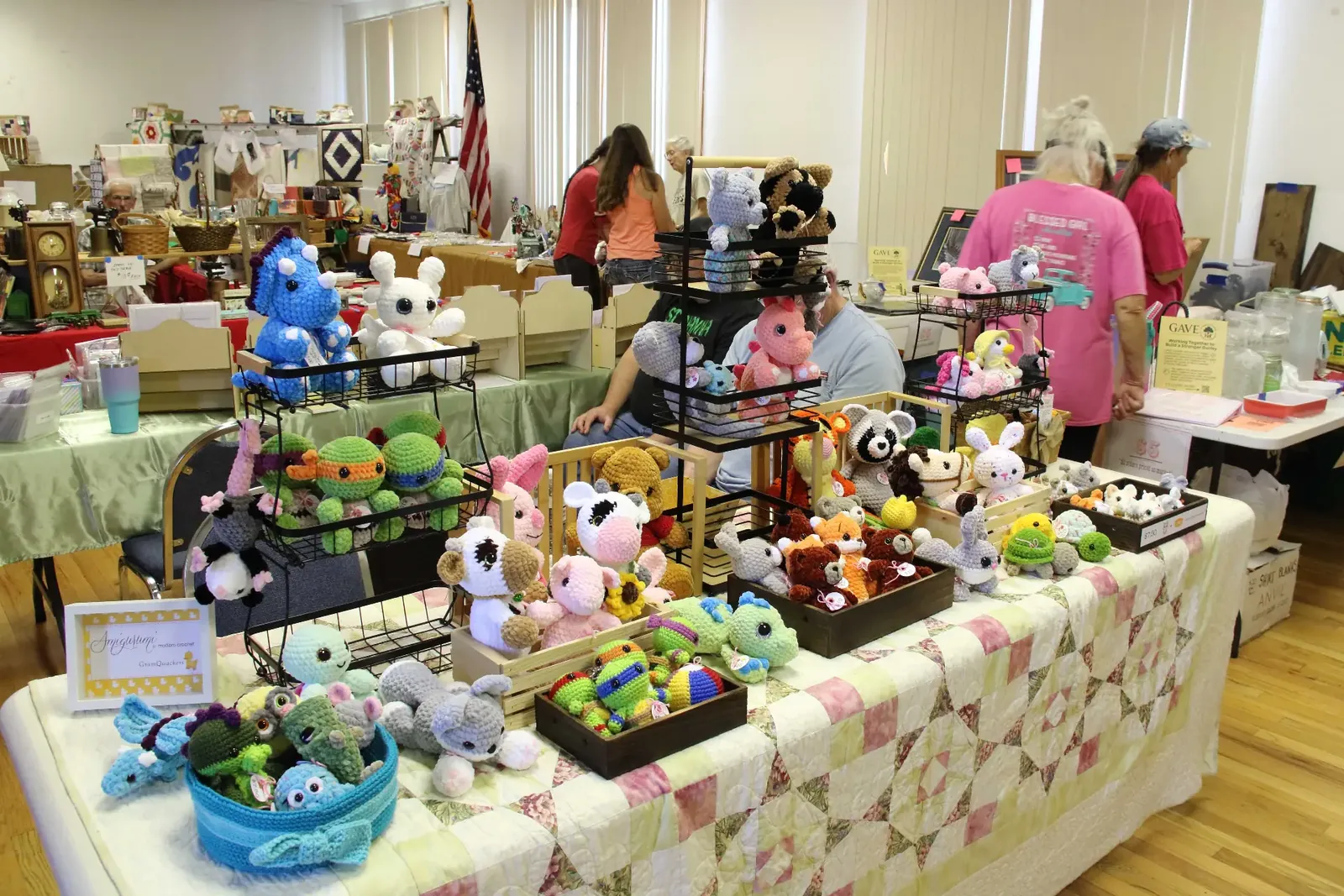Table covered with a patchwork quilt displaying colorful hand-crocheted animal plushies at an indoor craft fair