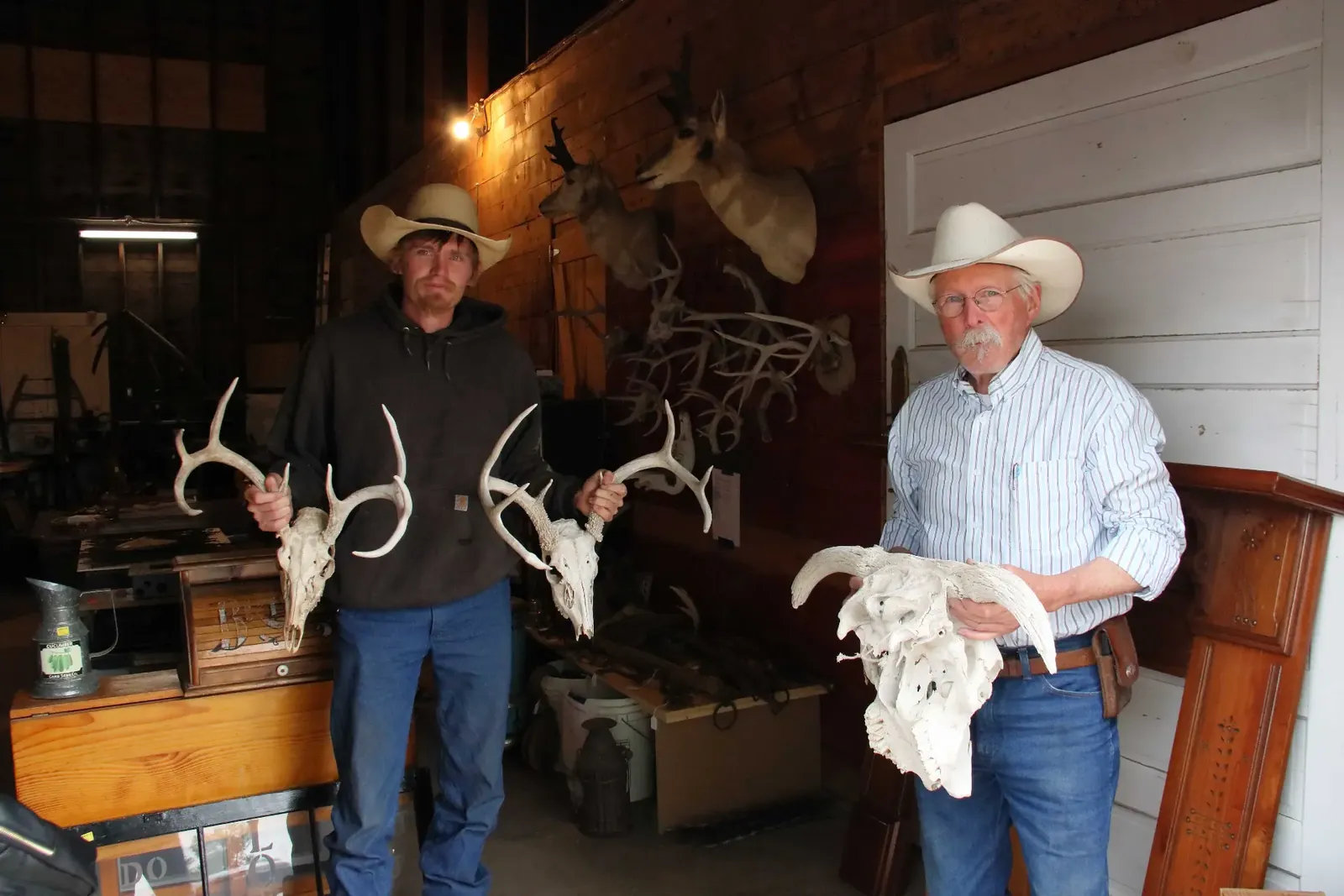 Two men in cowboy hats holding antlers and an animal skull in a workshop